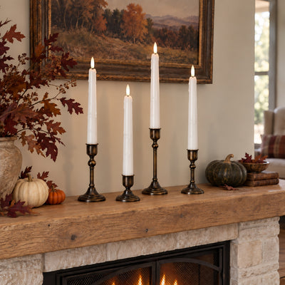 Four white taper candles in dark metal candlesticks displayed on a rustic wooden fireplace mantel in a cozy living room setting.