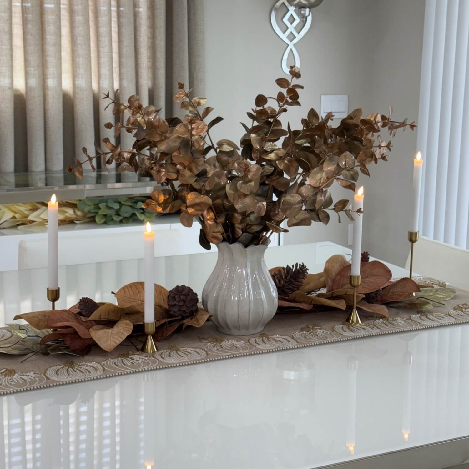 Decorative table setting with a vase of dried leaves, candles, and a runner on a white table.