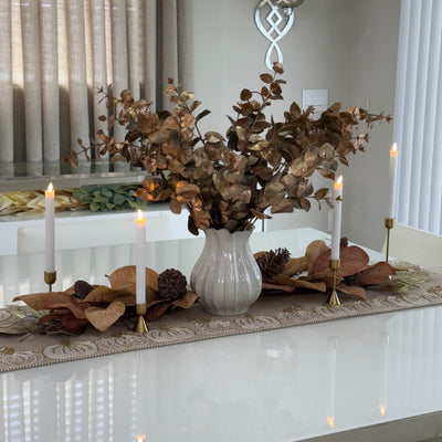 Decorative table setting with a vase of dried leaves, candles, and a runner on a white table.